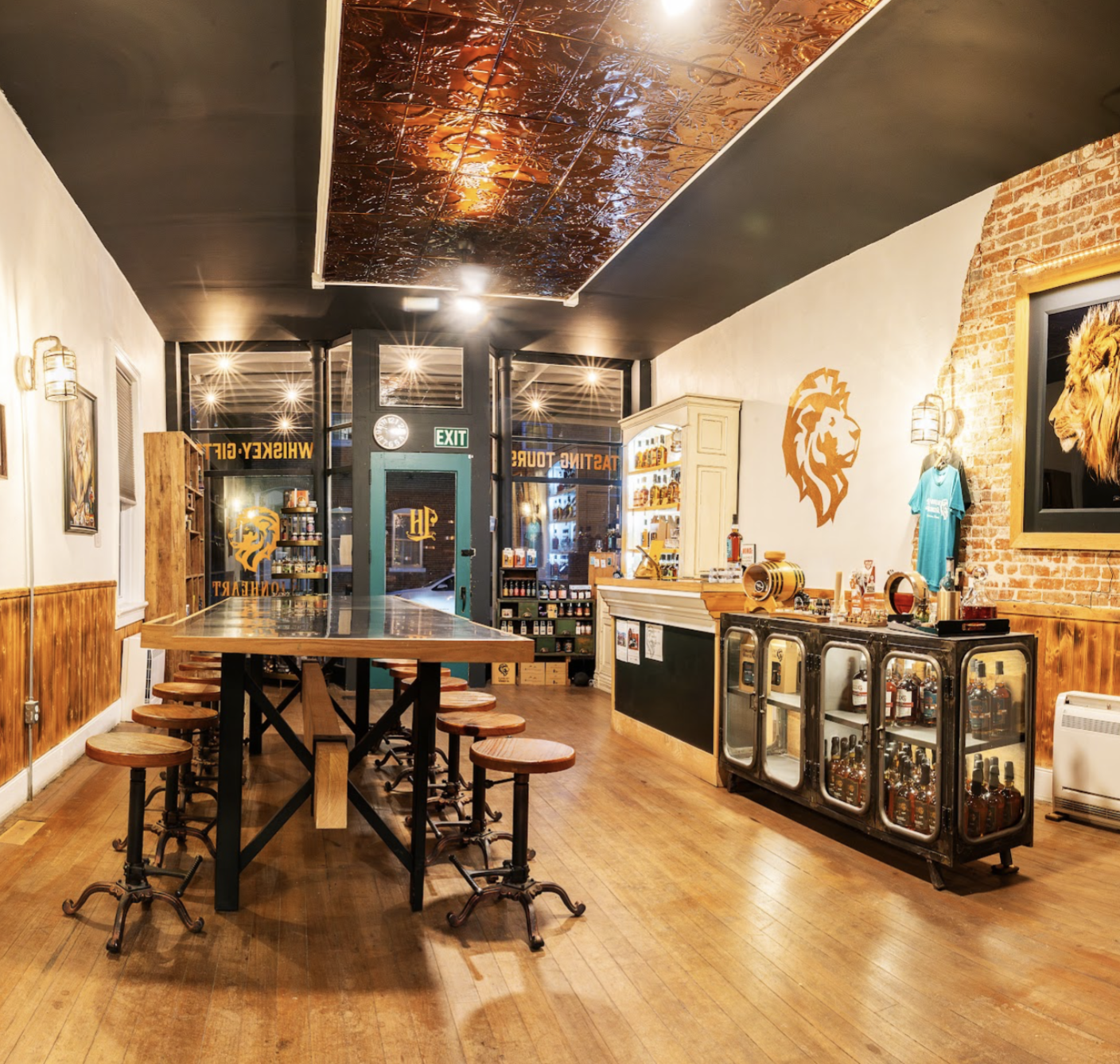 A brand ambassador pours whiskey from a Lionheart bottle into a tasting flight of small glasses arranged on a wooden tasting board. Guests sit at the community table family style in the background, with additional glassware, whiskey balls, and a decorative sculpture visible along the table.