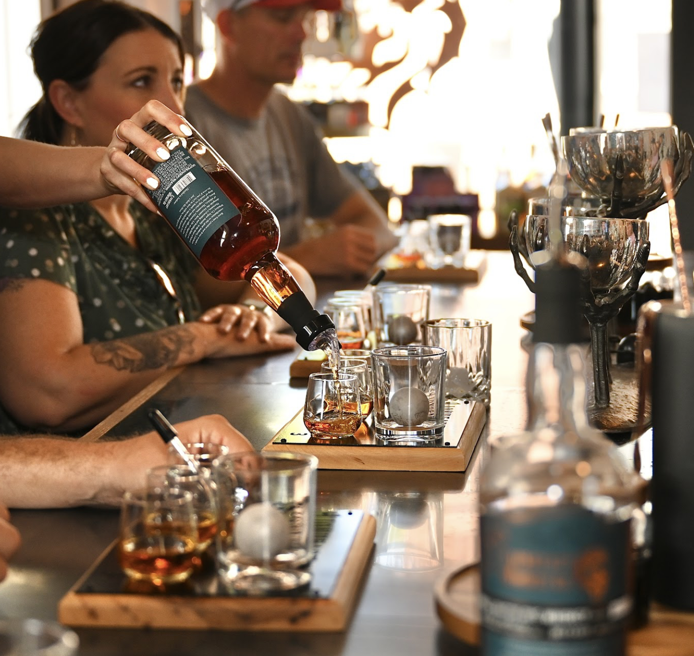 A brand ambassador pours whiskey from a Lionheart bottle into a tasting flight of small glasses arranged on a wooden tasting board. Guests sit at the community table family style in the background, with additional glassware, whiskey balls, and a decorative sculpture visible along the table.