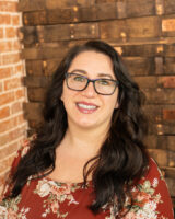 Brandi Lambright's headshot inside the Lionheart tasting room. Her black hair flowing over her shoulders and unmistakeable dark rimmed glasses show her personality.