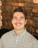 Ron Thomas smiles directly at the camera inside the Lionheart tasting room. He's wearing a plaid button down.